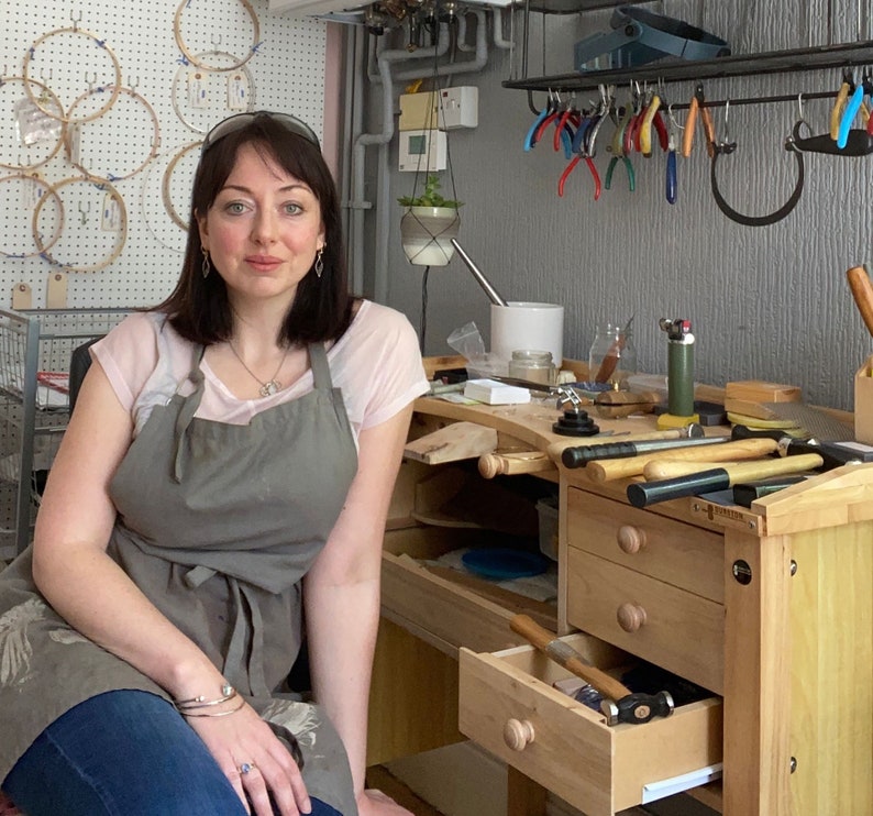 May include: A woman wearing a gray apron and a white shirt is standing in front of a workbench with tools and jewelry making supplies. The workbench has drawers and a wooden top.