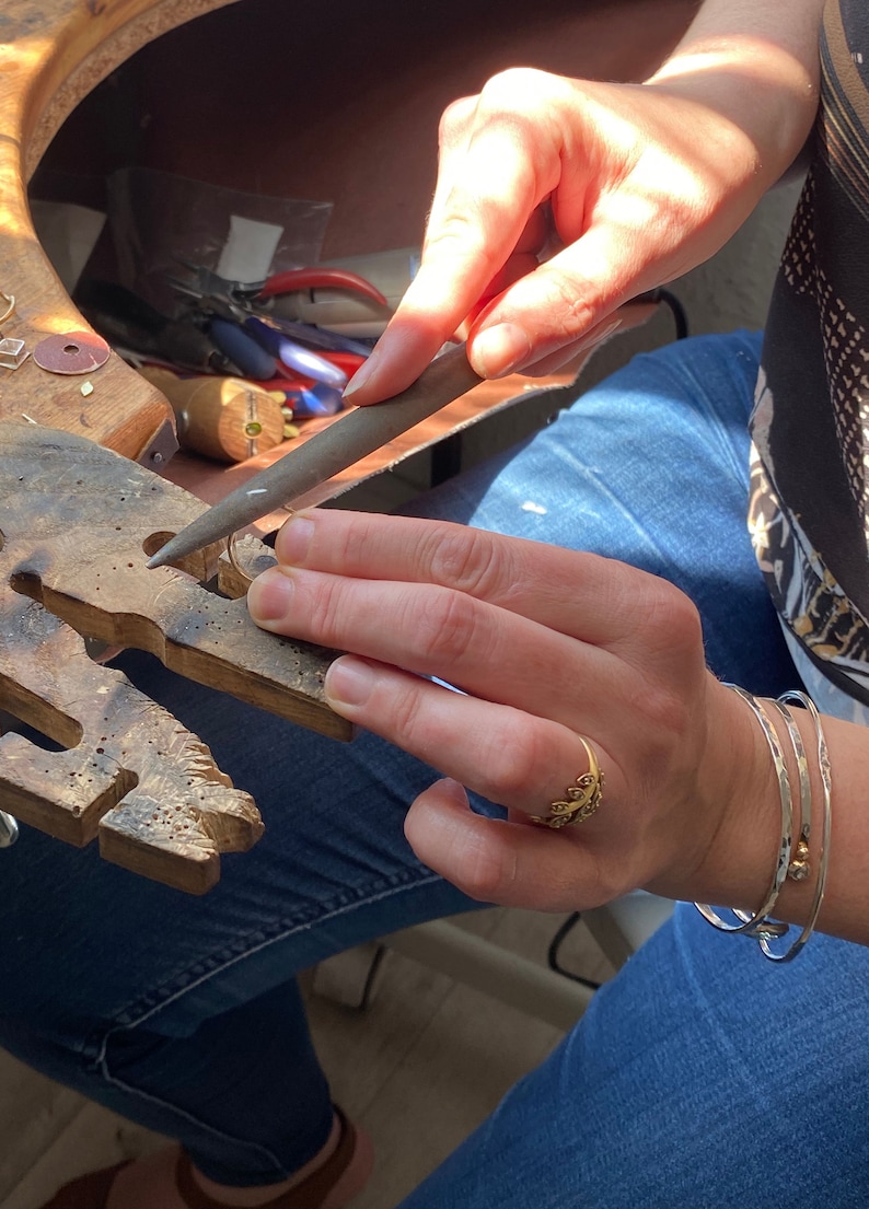 May include: A person is working on a piece of jewelry with a file. They are wearing a gold ring with a leaf design and two silver bracelets.