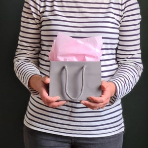 May include: A person holding a gray gift bag with a pink tissue paper lining. The bag has a gray rope handle.