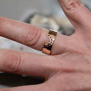 K&ouml;nnte beinhalten: Ein goldfarbener Ring mit einem einzigartigen, geometrischen Design. Der Ring ist mit kleinen, runden, roten Edelsteinen besetzt. Er wird an einem Finger getragen und unterstreicht seinen modernen und eleganten Stil. Das Design des Rings beinhaltet einen Ausschnitt.