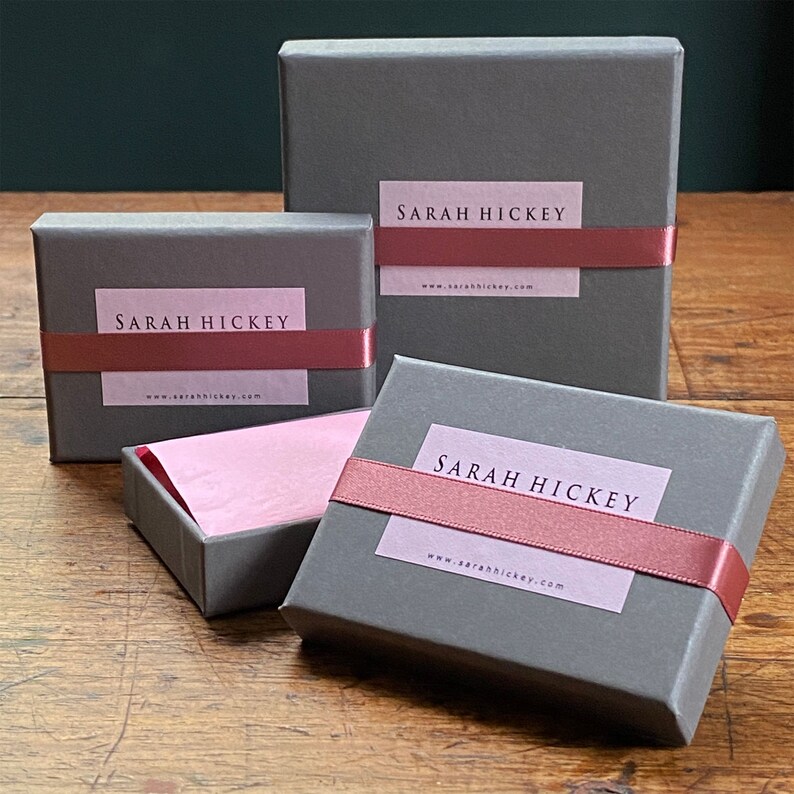 K&ouml;nnte beinhalten: Vier graue Geschenkboxen in verschiedenen Gr&ouml;&szlig;en, jede mit einem rosa Etikett und einem passenden Band. Die Etiketten tragen die Aufschrift "SARAH HICKEY" und enthalten eine Website-Adresse. Eine Box ist ge&ouml;ffnet und zeigt rosa Seidenpapier.
