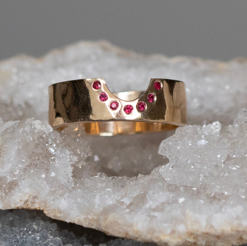 K&ouml;nnte beinhalten: Ein goldfarbener Ring mit einem geschwungenen Design, besetzt mit einer Reihe kleiner, runder, roter Edelsteine. Der Ring hat ein breites Band und eine einzigartige Form, wobei die Edelsteine entlang der oberen Kurve eingesetzt sind. Der Ring wird auf einer wei&szlig;en, strukturierten Oberfl&auml;che pr&auml;sentiert.