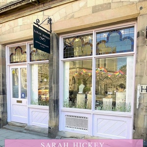 May include: A storefront with a pink facade and a sign that reads "Sarah Hickey Artisan Jewelry Est. 2013". The storefront has a large window with a rainbow flag banner hanging in the window. The storefront is located on a brick street.