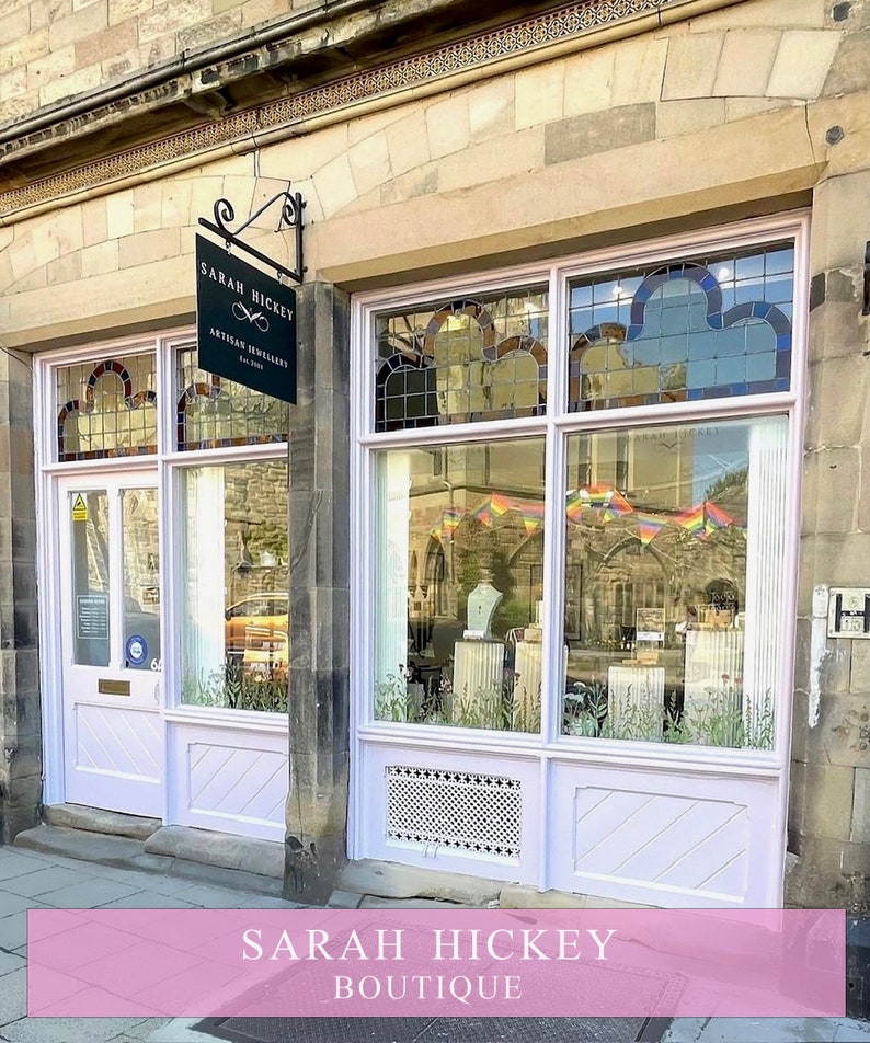 May include: A storefront with a pink facade and a sign that reads "Sarah Hickey Artisan Jewelry Est. 2013". The storefront has a large window with a rainbow flag banner hanging in the window. The storefront is located on a brick street.