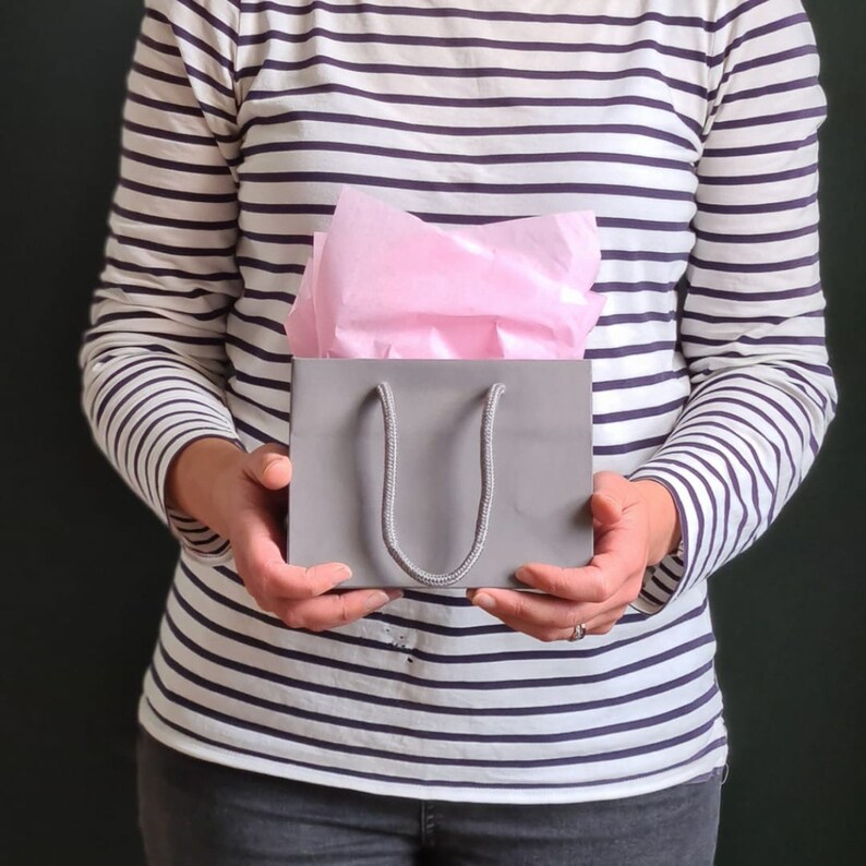 May include: A person holding a gray gift bag with a pink tissue paper lining. The bag has a gray rope handle.