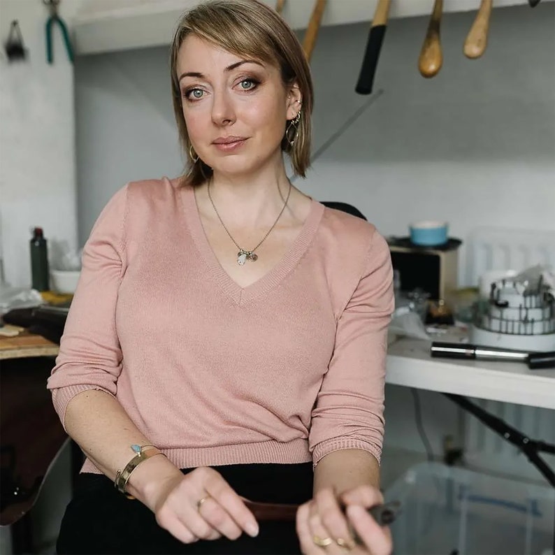 K&ouml;nnte beinhalten: Eine Frau tr&auml;gt einen hellrosa V-Ausschnitt-Pullover, Goldschmuck und eine Halskette mit Anh&auml;ngern. Die Frau befindet sich in einer Werkstatt, im Hintergrund sind Werkzeuge und Ger&auml;te zu sehen. Sie h&auml;lt ein Werkzeug.