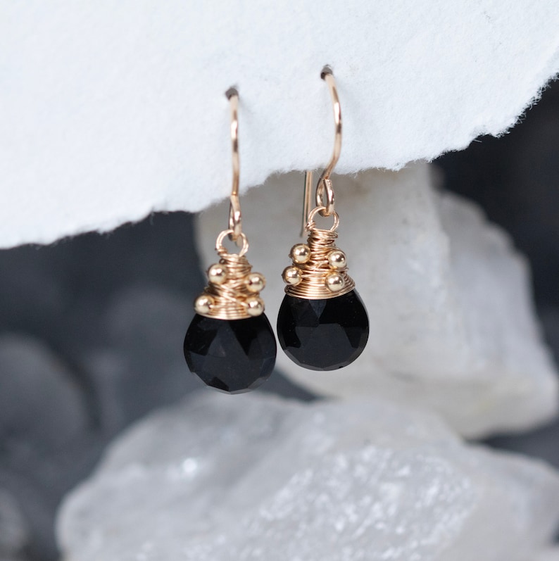 May include: A pair of gold earrings with black gemstone drops. The earrings are made with gold wire and have small gold beads around the top of the gemstone.