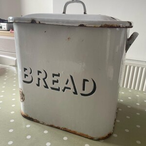 May include: White enamel bread bin with a black "BREAD" inscription. The bin has a hinged lid and a handle on top. The bin shows signs of wear and rust.