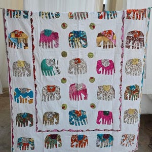 May include: White textile featuring a repeating pattern of colorful elephants. The elephants are in various colors and patterns, including pink, blue, and yellow. The border has a scalloped edge with a red and purple design. The textile is hanging.