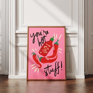 Pode incluir: Uma impressão artística rosa emoldurada com as palavras "you're hot stuff!" em escrita preta. A impressão apresenta ilustrações de pimentas vermelhas e uma garrafa de sriracha. A moldura é marrom.