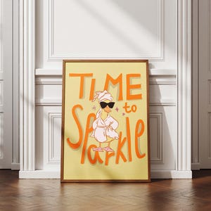 Könnte beinhalten: Ein gerahmter Kunstdruck mit den Worten "Time to Sparkle" in orangefarbenen Buchstaben auf gelbem Hintergrund. Das Kunstwerk zeigt eine Cartoon-Ente mit Sonnenbrille, einem Handtuch auf dem Kopf und einem Bademantel. Der Druck wird auf einem Holzboden präsentiert.