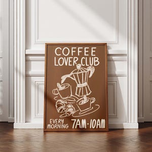 May include: A framed brown sign with white chalk-style illustrations of coffee cups, a coffee pot, and croissants. The text "COFFEE LOVER CLUB" and "EVERY MORNING 7AM-10AM" are written on the sign. The sign is displayed against a white wall and a wooden floor.