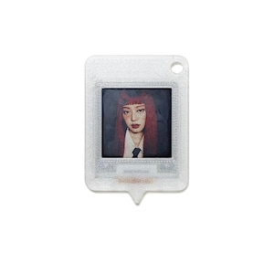 Op de afbeelding: Een doorzichtige, rechthoekige sleutelhanger met een portret van een persoon met rood haar. Het portret is omlijst in de sleutelhanger, die een klein gaatje aan de bovenkant en een puntige onderkant heeft. De achtergrond is wit.