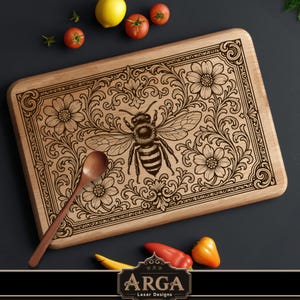 Könnte beinhalten: Holzschneidebrett mit detailliertem Laser-Gravur-Design mit einer Biene und einem Blumenmuster. Inklusive eines Holzkochlöffels. Das Brett ist rechteckig mit abgerundeten Ecken. Das Bild zeigt auch Tomaten, eine Zitrone und Paprika.