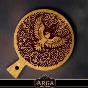 May include: A circular wooden serving board with a handle, displaying a laser-etched design of a golden eagle with outstretched wings, set against swirling patterns on a burgundy backdrop. The board has a natural wood grain finish and a small hole in the handle.