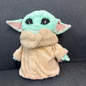 K&ouml;nnte beinhalten: Eine Pl&uuml;schpuppe einer Figur mit gro&szlig;en schwarzen Augen, gr&uuml;ner Haut und gro&szlig;en Ohren. Die Puppe tr&auml;gt einen beigefarbenen Mantel mit flauschigem Kragen und Manschetten. Die Puppe sitzt auf einer dunkelgrauen Oberfl&auml;che.