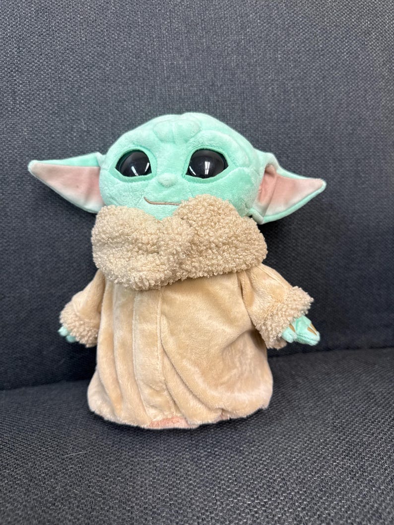 K&ouml;nnte beinhalten: Eine Pl&uuml;schpuppe einer Figur mit gro&szlig;en schwarzen Augen, gr&uuml;ner Haut und gro&szlig;en Ohren. Die Puppe tr&auml;gt einen beigefarbenen Mantel mit flauschigem Kragen und Manschetten. Die Puppe sitzt auf einer dunkelgrauen Oberfl&auml;che.