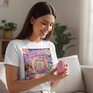 Peut inclure: T-shirt blanc avec un motif Tamagotchi color&eacute;, avec le texte "Tamagotchi". Un appareil Tamagotchi rose est tenu. Le design comprend des personnages de dessins anim&eacute;s et un th&egrave;me d'animal de compagnie num&eacute;rique.