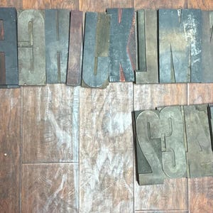 May include: A collection of vintage wooden letterpress printing blocks, including letters and numbers. The blocks are various sizes and colors, with visible wear and tear. The arrangement is on a wooden surface, showcasing the aged appearance of the printing tools.