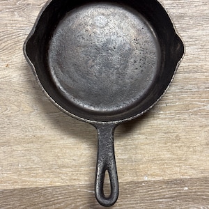 May include: A well-seasoned, cast iron skillet with a single handle and a rounded cooking surface. The pan is dark gray with a textured surface, showing signs of use. The handle has a hole for hanging. Ideal for cooking.