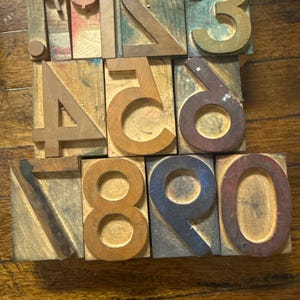 May include: A collection of vintage wooden printing blocks featuring numbers and a dollar sign. The blocks are various sizes and colors, including brown, green, and blue. The numbers range from 0 to 9, with a dollar sign included.