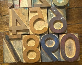 May include: A collection of vintage wooden printing blocks featuring numbers and a dollar sign. The blocks are various sizes and colors, including brown, green, and blue. The numbers range from 0 to 9, with a dollar sign included.