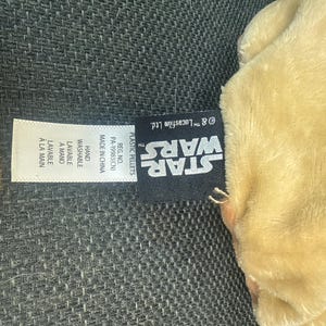 K&ouml;nnte beinhalten: Ein hellbraunes Pl&uuml;schtier mit einem Star Wars-Etikett. Das Etikett enth&auml;lt das Star Wars-Logo, Waschanweisungen und den Text "Made in China". Das Spielzeug scheint aus weichem Material zu sein.