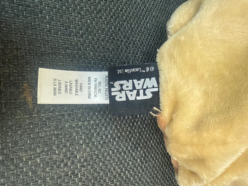 K&ouml;nnte beinhalten: Ein hellbraunes Pl&uuml;schtier mit einem Star Wars-Etikett. Das Etikett enth&auml;lt das Star Wars-Logo, Waschanweisungen und den Text "Made in China". Das Spielzeug scheint aus weichem Material zu sein.