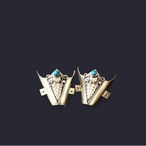 May include: A pair of gold-coloured metal decorative pieces, each featuring a turquoise stone and leaf design. The pieces have an ornate design and are set against a dark background.