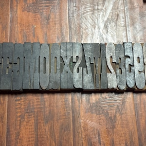 May include: A horizontal row of individual rectangular wooden letterpress blocks arranged side by side. The blocks feature reversed, raised characters including letters, numbers, and symbols, showing a weathered, dark brown wood grain texture.