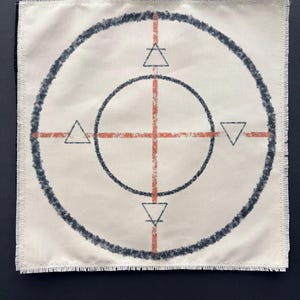 May include: A square off-white fabric patch with frayed edges featuring a dark gray circular design. An orange cross intersects the circles, and four small triangles are positioned at the cardinal points, with horizontal lines through the top and bottom triangles.