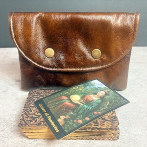 May include: A dark brown leather pouch with a curved flap and two brass-colored snap buttons. In front of the pouch sits a deck of cards with a decorative brown pattern on the edges, topped with a single tarot card labeled Queen of Pentacles.