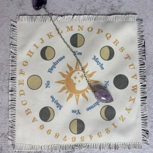 May include: A divination pendulum with a purple crystal and chain hangs over a cream-colored Ouija board cloth. The cloth features letters, numbers, moon phases, and the words "Yes," "No," and "Maybe."