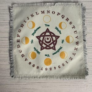 May include: A square woven fabric mat with fringed edges featuring a circular alphabet and number border. The center displays a maroon pentagram with a crescent moon and star, surrounded by yellow moon phases and the words Yes, No, Maybe, Rephrase.