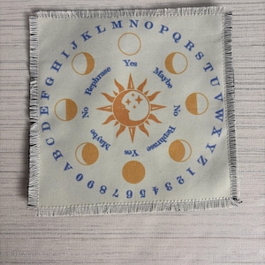 May include: A square woven fabric mat with fringed edges featuring a circular design. The center displays a sun and moon graphic with stars. Surrounding this are moon phases, the alphabet, numbers 0-9, and the words Yes, No, Maybe, and Rephrase.