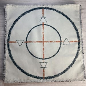 May include: Square, off-white fabric with a fringed edge. A dark blue, textured circle surrounds a lighter circle. Inside, a cross of orange lines intersects. Triangles point inward at the cardinal directions.