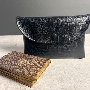 May include: A black pouch with a textured, embossed crocodile-skin pattern and a single snap closure, positioned behind a rectangular deck of cards featuring a dark brown and gold floral pattern with metallic gold-toned edges.