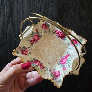 May include: A square, cream-coloured ceramic serving dish with a gold handle and trim. The dish is decorated with pink roses and green leaves. The centre of the dish has a gold floral pattern.