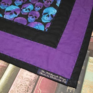 May include: A quilt featuring a central panel of blue and purple skulls on a black background, surrounded by a solid black border and a wide purple border. The fabric includes a label reading The Patchwork Home Made in Wales by Fil.