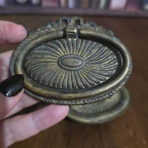May include: Antique brass drawer pulls with an oval design. The pulls have a ring handle and a decorative sunburst pattern. The brass has a weathered patina, suggesting age and use. The pulls are likely for a cabinet or drawer.