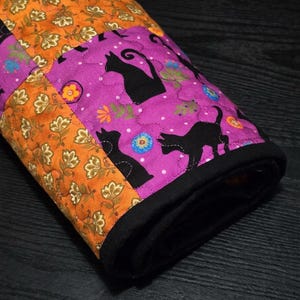 May include: A quilted fabric roll featuring alternating panels of orange fabric with a gold floral pattern and purple fabric with a black cat and colourful flower print. The roll is finished with a solid black fabric binding along the edge.