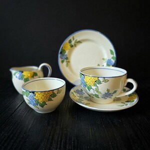 May include: A vintage tea set comprising a cup, saucer, bowl, creamer, and plate. The cream-coloured pieces are decorated with yellow and blue floral patterns, green leaves, and a blue trim. The set is displayed on a dark wooden surface.