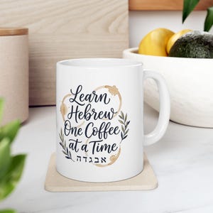 Peut inclure: Un mug en céramique blanche avec le texte noir Learn Hebrew One Coffee at a Time et des caractères hébreux. Le motif comprend des taches de café marron clair en forme d'anneaux et des illustrations de feuilles vertes.