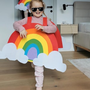 Puede incluir: Un niño o niña lleva un disfraz de arcoíris con nubes. El disfraz está hecho de fieltro y tiene un arcoíris rojo, naranja, amarillo, verde, azul, añil y violeta con nubes blancas.