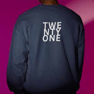 Könnte beinhalten: Ein marineblaues Sweatshirt mit dem Text "TWENTY ONE" in weißen Blockbuchstaben auf dem Rücken. Das Sweatshirt ist einfarbig und hat lange Ärmel. Der Hintergrund ist ein leuchtendes Magenta.