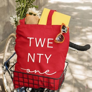 Op de afbeelding: Een rode draagtas met de witte tekst "TWENTY one", gevuld met een geel boek, zonnebril en bloemen, staat in een zwarte fietsmand. De fiets heeft een blauw frame en staat op een verharde ondergrond.