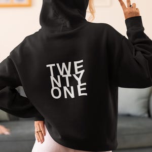 Könnte beinhalten: Schwarzer Kapuzenpullover mit dem weißen Aufdruck "TWENTY ONE". Der Hoodie hat eine Kapuze und lange Ärmel. Die Person trägt einen Ring und zeigt das Friedenszeichen mit den Fingern.