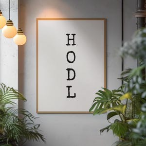May include: A framed print displaying the word "HODL" in black, bold letters. The print has a white background and a light brown frame. The image is set against a grey wall, with hanging lights and greenery in the foreground.