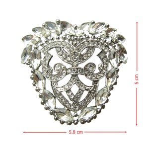 Op de afbeelding: Een zilveren hartvormige broche met een decoratief ontwerp en heldere strass steentjes. De broche is 5,8 cm breed en 5 cm hoog.