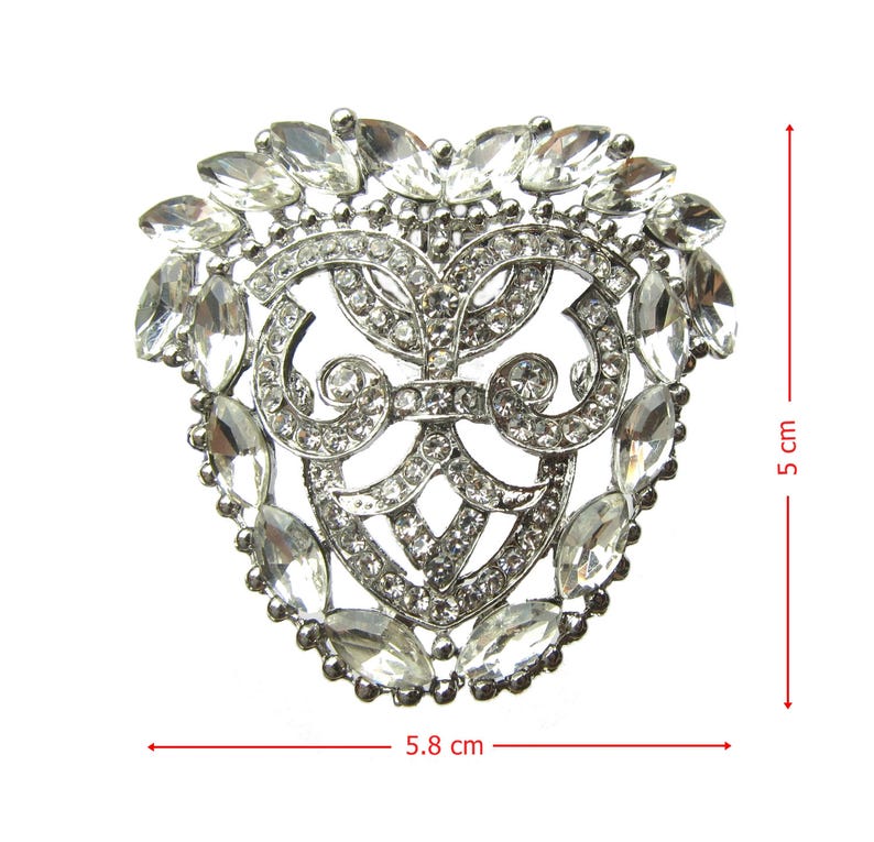 Op de afbeelding: Een zilveren hartvormige broche met een decoratief ontwerp en heldere strass steentjes. De broche is 5,8 cm breed en 5 cm hoog.